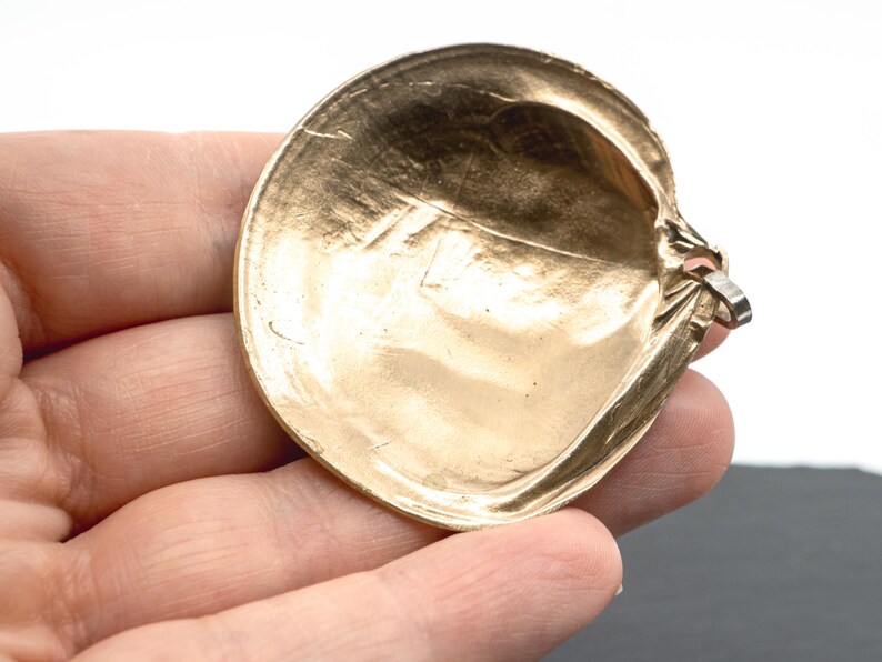 May include: A gold-toned metal pendant shaped like a rounded seashell. The surface has a smooth, reflective finish with subtle indentations and a metallic sheen. A small silver-colored metal loop is attached to the side for hanging or attachment.