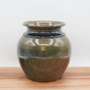 May include: A handcrafted ceramic vase with a rounded body and a slightly flared rim. The vase features a blend of earthy tones, including brown, green, and blue, with a glossy finish. The vase is sitting on a wooden surface.