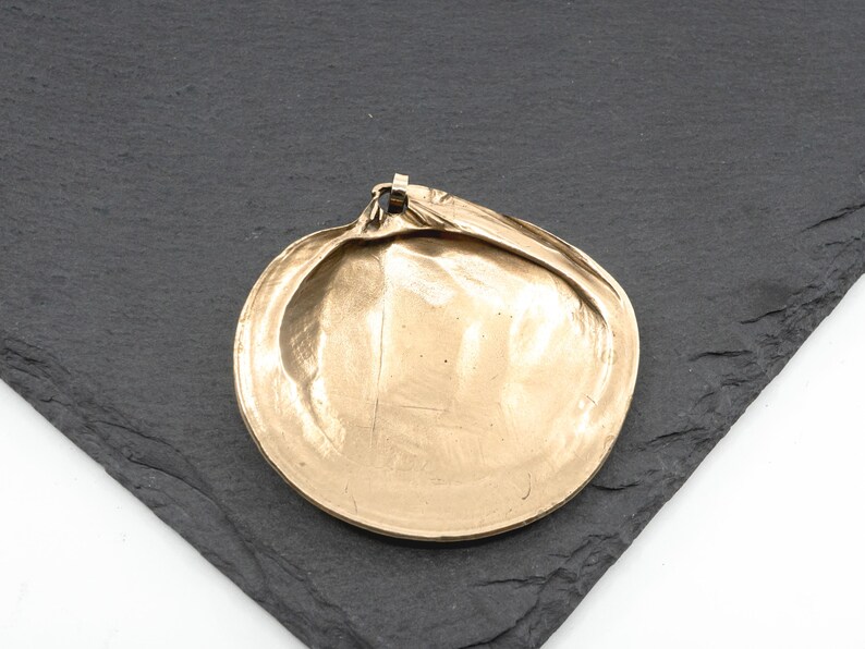 May include: A gold-toned metal pendant shaped like a clam shell. The surface has a smooth, reflective finish with subtle ridges and a small loop at the top for attachment. The object is circular in form with a slightly irregular, organic edge.