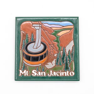 May include: A square ceramic tile featuring a relief illustration of the Mt. San Jacinto aerial tramway. The design includes an orange tram car, green pine trees, and brown mountain cliffs against a blue sky, with the text Mt. San Jacinto below.