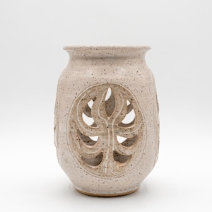 May include: A speckled beige ceramic vase with a leaf design cut out of the front. The vase has a wide opening and a rounded body. The leaf design is centered on the front of the vase.