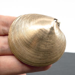 May include: A gold-toned metal pendant shaped like a clam shell, featuring a detailed, concentric ridged texture across its surface. The pendant has a small loop at the top for attachment, showcasing a metallic finish with subtle light reflections.