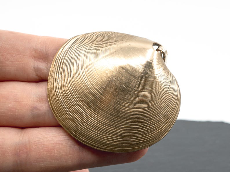 May include: A gold-toned metal pendant shaped like a clam shell, featuring a detailed, concentric ridged texture across its surface. The pendant has a small loop at the top for attachment, showcasing a metallic finish with subtle light reflections.