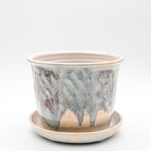 May include: A ceramic planter with a matching saucer. The planter has a cylindrical shape with a wide opening and a light beige rim. The exterior features a blend of blue, purple, and beige glaze, creating a dripped effect. The saucer is a similar color.