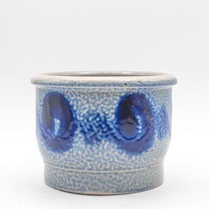 May include: A small, round, ceramic planter with a textured, speckled blue-gray exterior. The planter features a dark blue, stylized floral design around its circumference. The rim and base are a lighter shade of blue-gray.