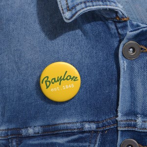 May include: A yellow button pin with the word "Baylor" in green script and the text "est. 1845" in white. The pin is affixed to a blue denim jacket.