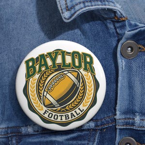 May include: A white button with the words "BAYLOR FOOTBALL" in green and gold. The button features a football graphic in the center, surrounded by a gold wreath design. The button is set against a blue denim background.