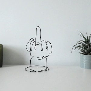 Könnte beinhalten: Eine schwarze Drahtskulptur einer Hand, die den Mittelfinger zeigt. Eine grüne Kerze in einem Milchglasbehälter und eine kleine Sukkulentenpflanze in einem hellblauen Topf befinden sich ebenfalls auf der weißen Oberfläche.