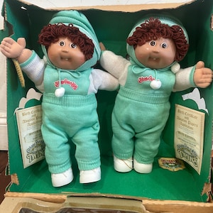 以下が含まれることがあります： 緑色の箱に入った2体のキャベツ人形。人形は茶色の糸の髪、色白の肌をしており、白のアクセントが付いたお揃いのミントグリーンの衣装を着ています。箱には出生証明書と養子縁組書類が含まれています。人形には「Twins」とラベルが付けられています。