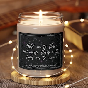 Puede incluir: Una vela marrón con una etiqueta negra que dice "Hold on to the memories, they will hold on to you." La etiqueta también tiene una pequeña estrella blanca y el texto "PLEASE DON'T EVER BECOME A STRANGER".