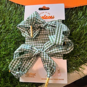 May include: Green and white gingham patterned shoelaces with gold-colored aglets. The shoelaces are tied into bows and are displayed on a white card with the text "STEP IT UP laces" and "the darling effect."