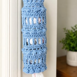 May include: A light blue, handmade crochet bag dispenser hanging on a white wall. The cylindrical holder features an openwork design, with a bag peeking out from the bottom. The dispenser is designed to hold plastic grocery bags.