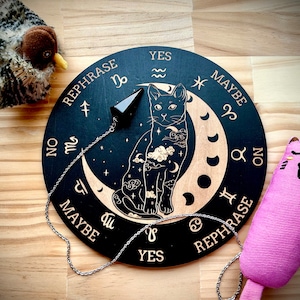 May include: A black wooden divination board with a cat and moon design, featuring the words "YES", "NO", "MAYBE", and "REPHRASE". A black crystal pendulum hangs over the board. Decorative cat toys are on either side.