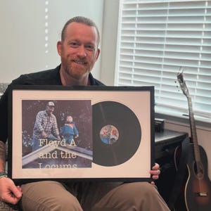 May include: Framed vintage vinyl record and photograph. The photograph features two people on a rooftop, with the text "Floyd A and the Locums" below. The vinyl record is black with a colorful label. The frame is black with a white mat.