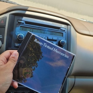 May include: A compact disc with a blue and white sky and a tree reflected in the disc. The title of the CD is "Rose-Tinted Memories".