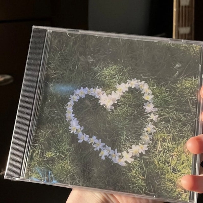 May include: A CD case with a photo of a heart shape made of white flowers on a green grassy background.
