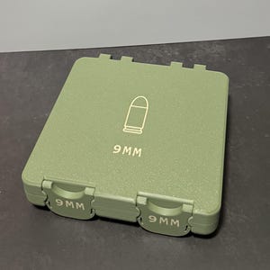 Può includere: Una custodia rettangolare verde oliva con coperchio incernierato. Il coperchio presenta un contorno bianco di un proiettile e la scritta "9MM". La custodia ha due chiusure, ciascuna con la scritta "9MM".