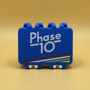 Könnte beinhalten: Eine blaue Phase 10 Kartenspielhülle mit weißem Text und bunten Streifen. Die Hülle hat eine rechteckige Form mit gewölbter Ober- und Unterseite. Der Text "Phase 10" ist prominent auf der Vorderseite abgebildet.