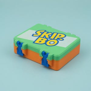 May include: A colorful SKIP BO game box. The box is rectangular with a green top, orange base, and white center panel with the game's name in blue and yellow. Blue clasps secure the box.