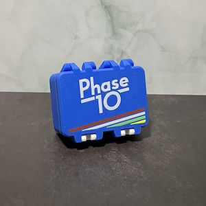 May include: A blue Phase 10 game case with white text and a colorful stripe design. The case has a rectangular shape with rounded edges and a handle on top. The game case is sitting on a dark surface.