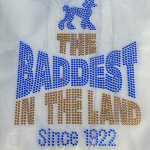 May include: A white t-shirt with a blue rhinestone design that says "The Baddest In The Land Since 1922". A blue rhinestone poodle is above the text.
