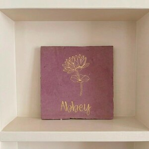 Op de afbeelding: Een vierkante, oudroze stenen tegel met een goudkleurige lijntekening van een lotusbloem gecentreerd boven de naam Abbey, geschreven in een cursief lettertype. De tegel heeft een mat, getextureerd oppervlak met een natuurlijke uitstraling.