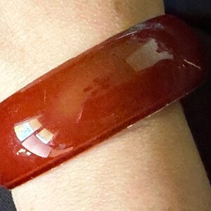 May include: A wide, smooth, deep red bangle bracelet with a glossy, reflective finish. The bracelet has a solid, uniform color and a rounded rectangular profile, encircling the wrist with a polished surface that catches light across its exterior.