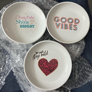 Könnte beinhalten: Drei weiße Keramikschalen mit inspirierenden Sprüchen. Eine Schale trägt die Aufschrift "Keep Calm & Shine Bright", eine andere "Good Vibes" und die dritte "be cool, stay wild" mit einem pinkfarbenen Leopardenherz.