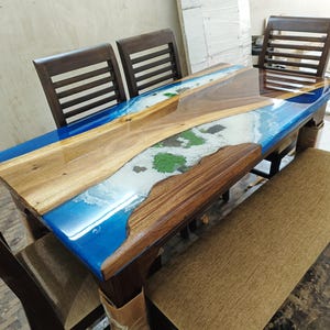 May include: A wooden dining table with a blue and white epoxy resin inlay that resembles a beach scene. The table is surrounded by four chairs and a bench.