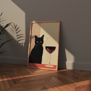 Op de afbeelding: Een ingelijst afdruk van een zwarte kat die naast een glas rode wijn zit. De kat kijkt recht naar de kijker. De achtergrond is lichtbeige.