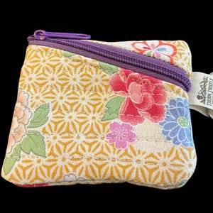 May include: A small square fabric coin pouch featuring a yellow geometric pattern with pink, red, and blue floral accents. It has a purple zipper closure and a silver metal lobster clasp keychain attached to a white tag labeled Thorn Thistle Thread.