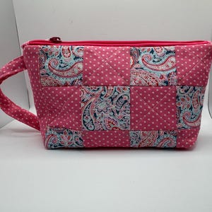 May include: A quilted zippered pouch featuring a patchwork design of alternating squares. The fabric alternates between a pink background with white polka dots and a multi-colored paisley print in shades of blue, red, and white. Includes a side handle.