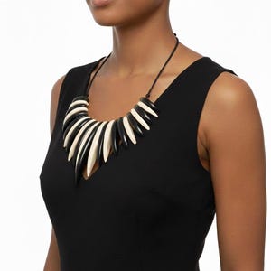 Peut inclure: Un collier imposant avec des éléments noirs et crème, incurvés et pointus en alternance. Le collier est monté sur un cordon noir. Le design crée un motif géométrique audacieux. Le collier est porté sur un haut sans manches noir, ajoutant un accent saisissant à une tenue.