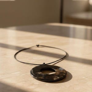 Puede incluir: Un collar con colgante circular negro y marrón con un agujero central. El colgante está suspendido de un fino cordón negro. El collar está sobre una superficie clara.