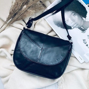 Puede incluir: Un bolso bandolera de cuero negro con cierre de solapa redondeada y una correa negra larga y ajustable. El bolso presenta una textura suave y un diseño estructurado con costuras visibles a lo largo de la solapa frontal y el cuerpo.