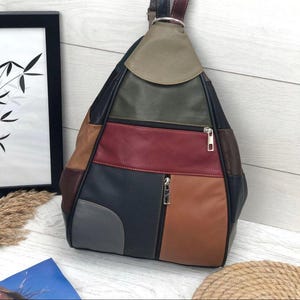May include: A triangular backpack constructed from multi-colored leather panels in shades of olive green, deep red, dark blue, tan, and grey. The bag features two horizontal zippered compartments with silver-toned metal pulls and adjustable straps.