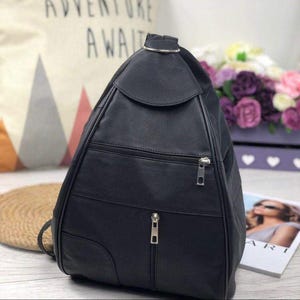 May include: A black teardrop-shaped backpack featuring a smooth finish, a top handle with a silver-toned metal ring, a horizontal zippered pocket across the middle, and a vertical zippered pocket extending from the center to the bottom edge.