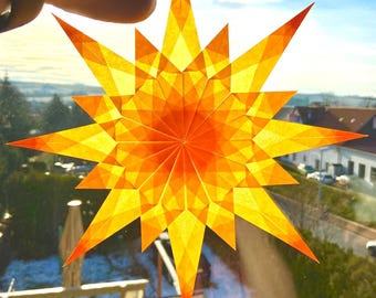 Könnte beinhalten: Ein mehrzackiger Sternschmuck aus durchscheinendem orangefarbenem und gelbem Papier mit einem geometrischen Faltmuster, das bei Gegenlicht durch Sonnenlicht einen strahlenden Effekt erzeugt und scharfe Spitzen zeigt.