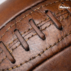 May include: Close-up of a brown leather football with stitching. The stitching is a lighter brown color and is visible throughout the image.