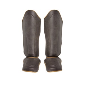 May include: A pair of black shin guards with gold trim. The shin guards are made of a padded material and have a contoured shape to fit the legs.