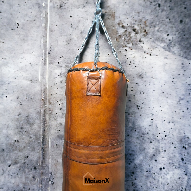 May include: A brown leather punching bag hanging from a silver chain against a gray concrete wall. The bag has black stitching and a leather strap with a metal loop.