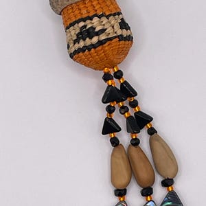 May include: Close-up of a handmade acorn-shaped pendant. The pendant features an orange woven body with black and cream-colored accents, a tan cap, and beaded strands with black triangles, wooden beads, and iridescent shell pendants.