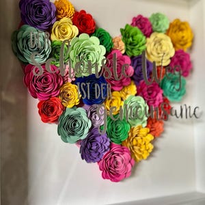Könnte beinhalten: Ein gerahmtes Blumenherz-Arrangement. Das Herz besteht aus bunten Papierblumen in Lila, Rosa, Gelb, Grün und Rot. Silberne Schrift lautet "Schönste Weg ist der gemeinsame". Der Rahmen ist weiß.