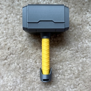 Personalized baby rattle shaped like Thor’s hammer, 3D printed with custom name engraving and multiple handle colors such as light blue, pink, black, brown, and yellow. A unique custom baby gift and fun 3D printed baby toy.