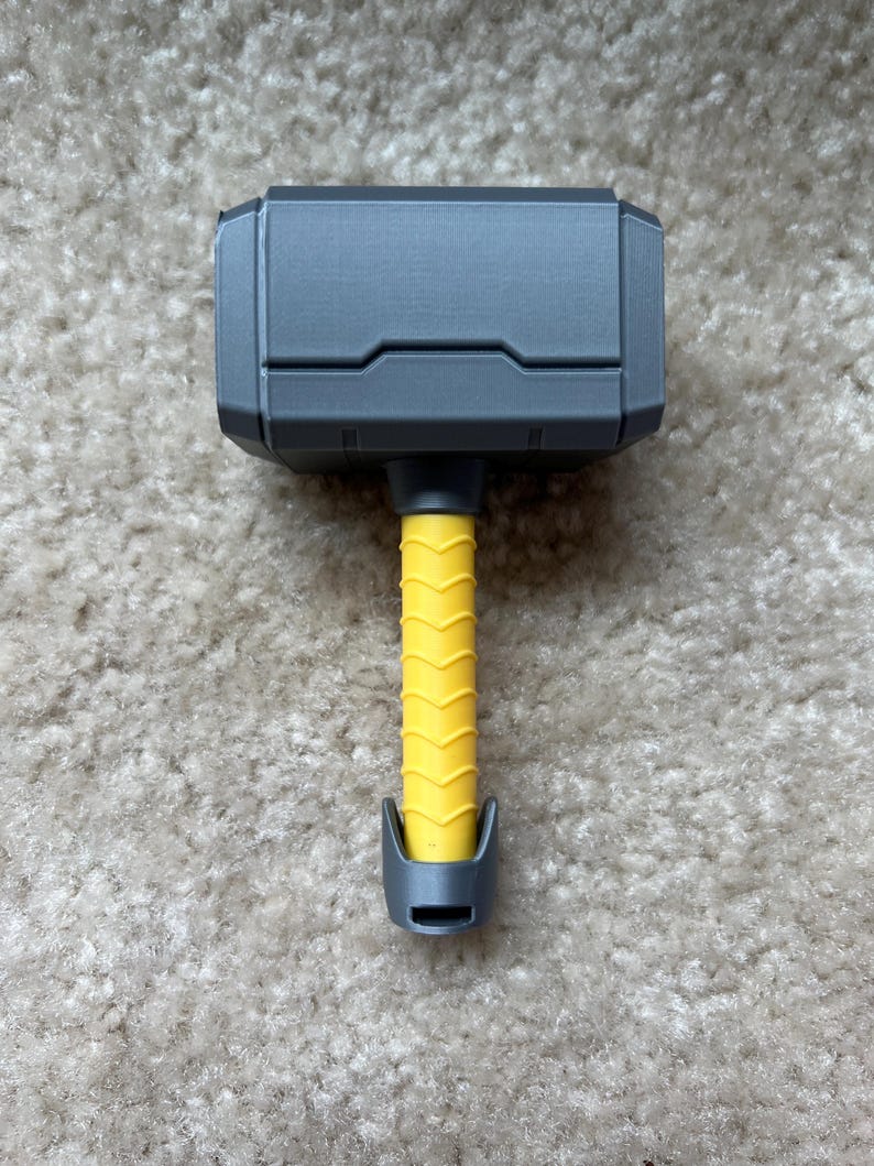 Personalized baby rattle shaped like Thor’s hammer, 3D printed with custom name engraving and multiple handle colors such as light blue, pink, black, brown, and yellow. A unique custom baby gift and fun 3D printed baby toy.