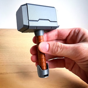 May include: A small, silver-colored hammer with a brown wooden handle. The hammer is a replica of Thor's hammer, Mjolnir, from the Marvel comics and movies.