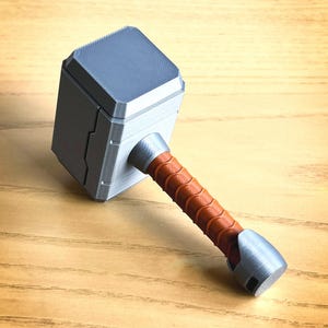 May include: A 3D printed silver Mjolnir hammer with a brown leather-wrapped handle. The hammer has a detailed design and a textured surface.
