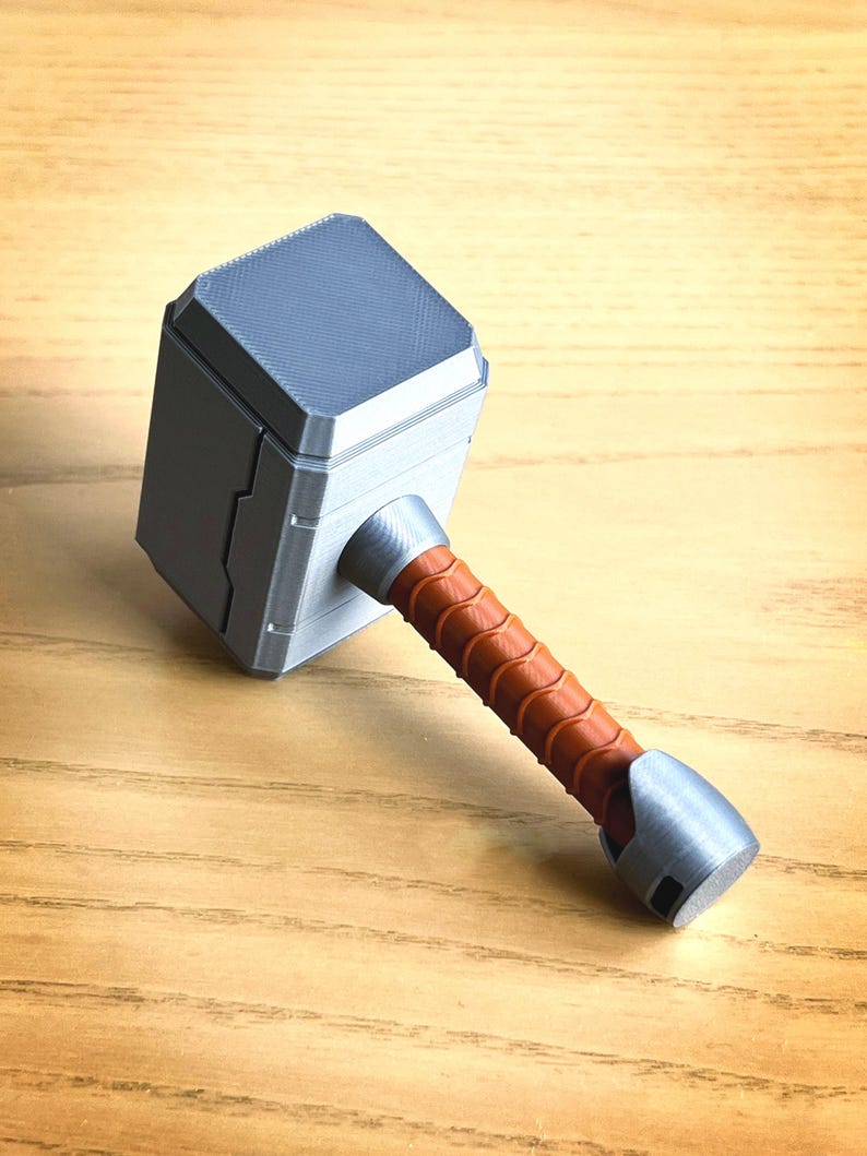 May include: A 3D printed silver Mjolnir hammer with a brown leather-wrapped handle. The hammer has a detailed design and a textured surface.