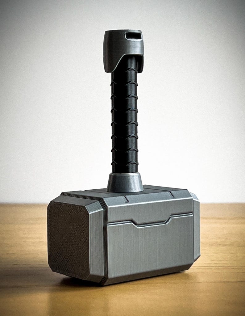 May include: A silver-colored, 3D printed replica of Thor's hammer, Mjolnir. The hammer has a black handle with a textured surface and a silver-colored head with a raised design.