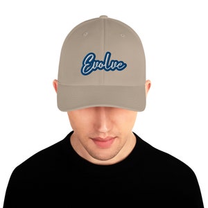 May include: A tan baseball cap with the word "Evolve" embroidered in blue script. The cap is a neutral color and has a curved brim. The person wearing the cap is wearing a black shirt.